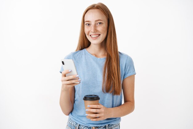 Pelirroja mujer linda, feliz con pecas sonriendo con taza de café y teléfono inteligente