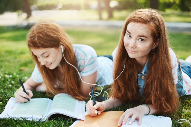 pelirrojo mirando con las cejas levantadas y una linda sonrisa, acostado en la hierba en el parque de la ciudad con su hermana, compartiendo auriculares para escuchar música juntos y haciendo la tarea. Concepto de estilo de vida y personas