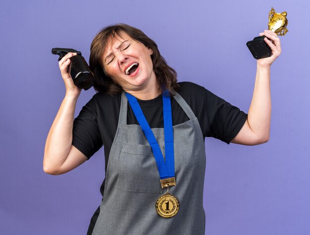 Peluquería femenina adulta disgustada en uniforme con medalla de oro alrededor del cuello sosteniendo una botella de spray y una copa ganadora aislada en la pared púrpura con espacio de copia