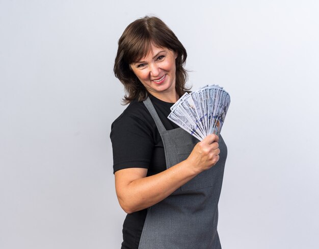 Peluquería femenina adulta sonriente en uniforme sosteniendo dinero aislado en la pared blanca con espacio de copia