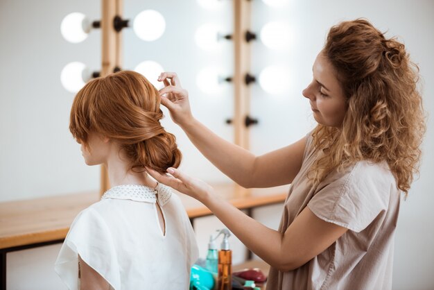 Peluquería femenina haciendo peinado a mujer pelirroja en salón de belleza