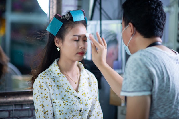 Peluquería femenina de pie, maquillaje facial y peinado a linda joven encantadora en salón de belleza