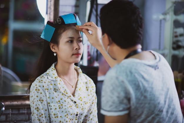 Peluquería femenina de pie, maquillaje facial y peinado a linda joven encantadora en salón de belleza