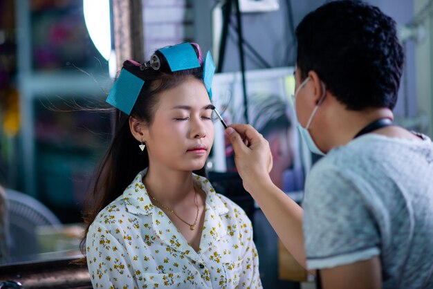 Peluquería femenina de pie, maquillaje facial y peinado a linda joven encantadora en salón de belleza