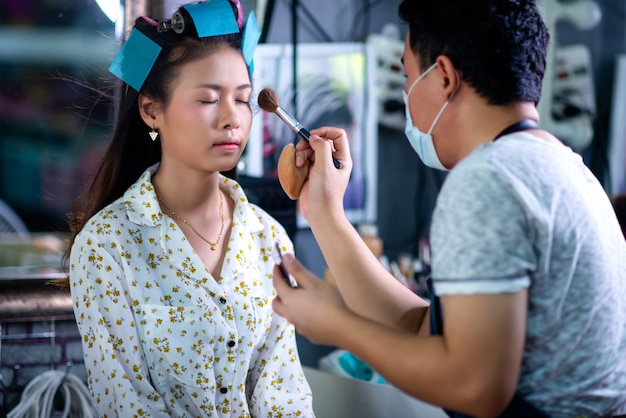 Peluquería femenina de pie, maquillaje facial y peinado a linda joven encantadora en salón de belleza