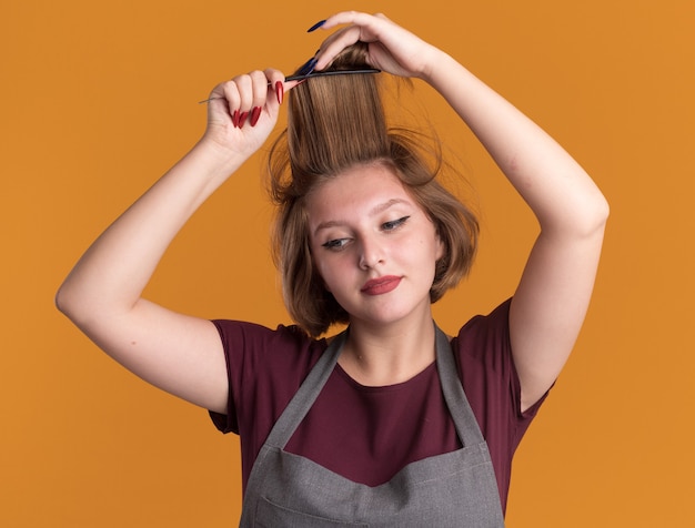 Peluquería joven hermosa en delantal peinándose el pelo mirando a un lado con una sonrisa en la cara de pie sobre la pared naranja