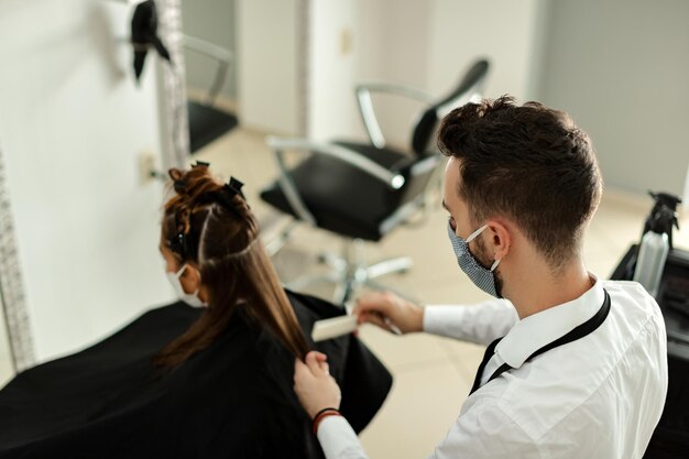 Peluquería con mascarilla protectora que trata el cabello de la mujer en el salón durante la epidemia de virus