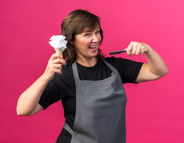 Peluquería mujer caucásica adulta alegre en uniforme sosteniendo navaja de afeitar y brocha de afeitar con espuma en la parte superior aislada sobre fondo rosa con espacio de copia