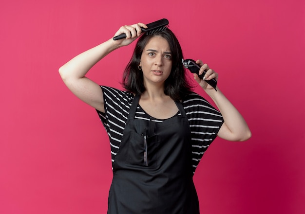Foto gratuita peluquería mujer hermosa joven en delantal con cortadora de cabello peinándose con cepillo de pie sobre la pared rosa