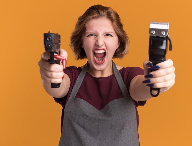 Peluquería de mujer hermosa joven en delantal con máquina recortadora apuntando con pistola gritando con expresión agresiva de pie sobre la pared naranja