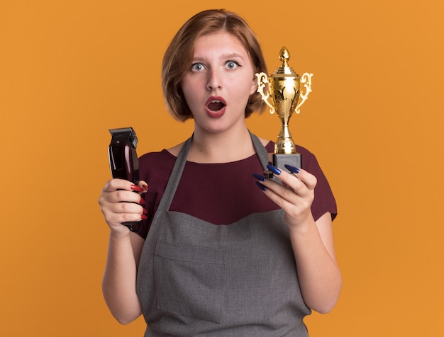 Peluquería de mujer hermosa joven en delantal con máquina recortadora y trofeo de oro mirando al frente sorprendido y asombrado de pie sobre la pared naranja