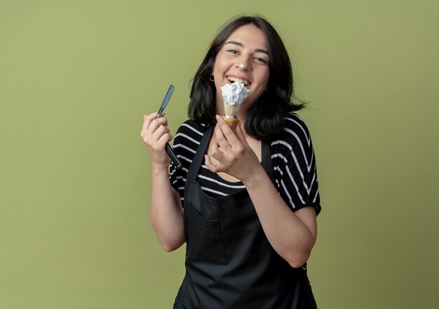 Peluquería mujer hermosa joven en delantal con maquinilla de afeitar y brocha de afeitar con espuma sonriendo alegremente de pie sobre la pared de luz