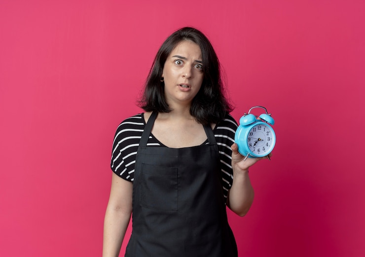 Foto gratuito peluquería mujer hermosa joven en delantal mostrando despertador preocupado de pie sobre la pared rosa