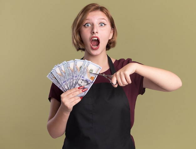 Peluquería de mujer hermosa joven en delantal mostrando dinero en efectivo tratando de cortar dinero con tijeras de pie emocional y emocionado sobre pared verde