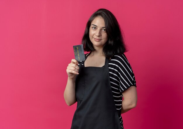 Peluquería mujer hermosa joven en delantal mostrando lookign tarjeta de crédito a la cámara sonriendo sobre rosa
