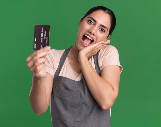 Peluquería de mujer hermosa joven en delantal mostrando tarjeta de crédito sonriente feliz y positiva de pie sobre la pared verde