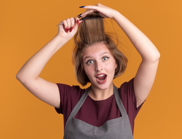 Foto gratuita peluquería de mujer hermosa joven en delantal peinándose el cabello mirando feliz y sorprendido de pie sobre la pared naranja
