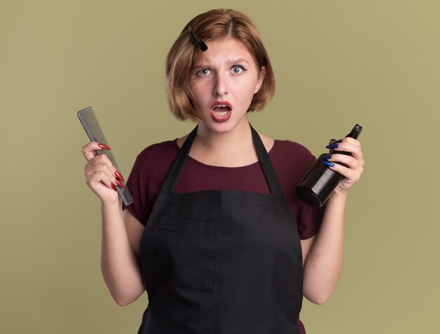 Peluquería de mujer hermosa joven en delantal con peine y botella de spray mirando al frente confundido y disgustado de pie sobre la pared verde