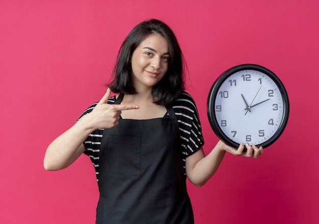 Peluquería mujer hermosa joven en delantal con reloj de pared apuntando con el dedo sonriendo de pie sobre la pared rosa