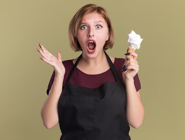 Peluquería de mujer hermosa joven en delantal sosteniendo brocha de afeitar con espuma de afeitar mirando al frente sorprendido de pie sobre la pared verde