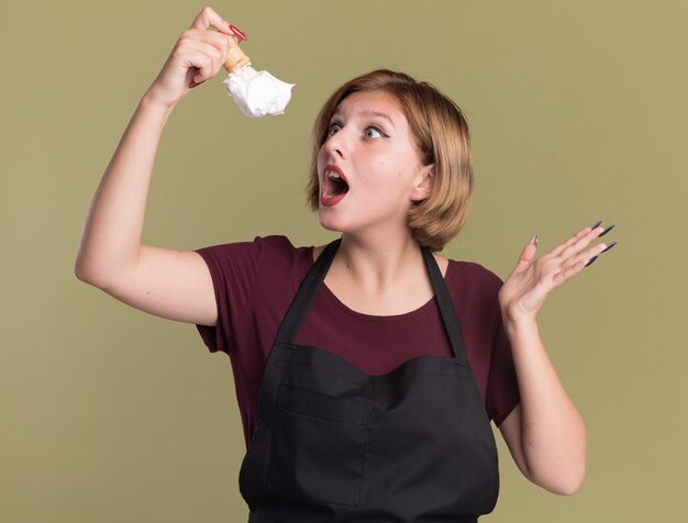 Peluquería de mujer hermosa joven en delantal sosteniendo brocha de afeitar con espuma de afeitar mirándolo asombrado parado sobre pared verde