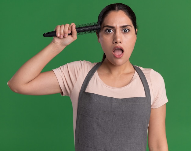 Peluquería de mujer hermosa joven en delantal sosteniendo cepillo de pelo sobre su cabeza mirando sorprendido y asombrado de pie sobre la pared verde