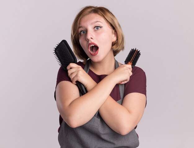 Peluquería de mujer hermosa joven en delantal sosteniendo cepillos de pelo cruzando las manos mirando sorprendido de pie sobre la pared blanca