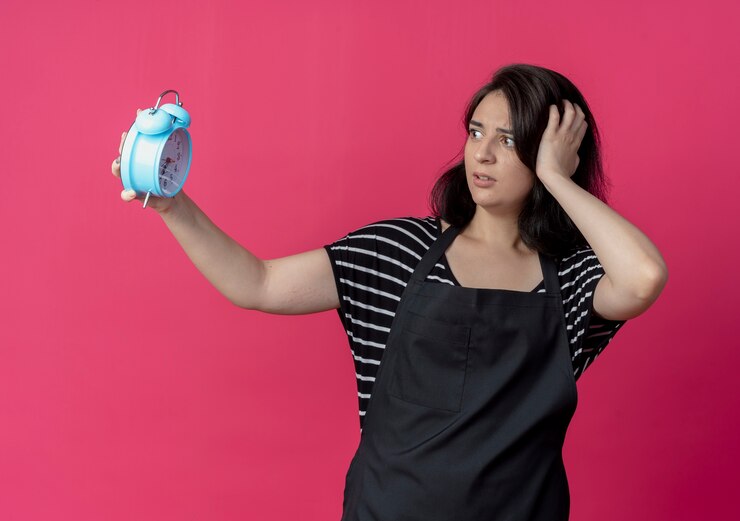 Foto gratuito peluquería mujer hermosa joven en delantal sosteniendo despertador mirándolo confundido y preocupado de pie sobre la pared rosa