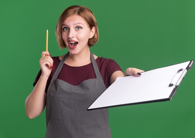 Peluquería de mujer hermosa joven en delantal sosteniendo lápiz mostrando portapapeles mirando al frente sorprendido y feliz de tener nueva idea de pie sobre la pared verde