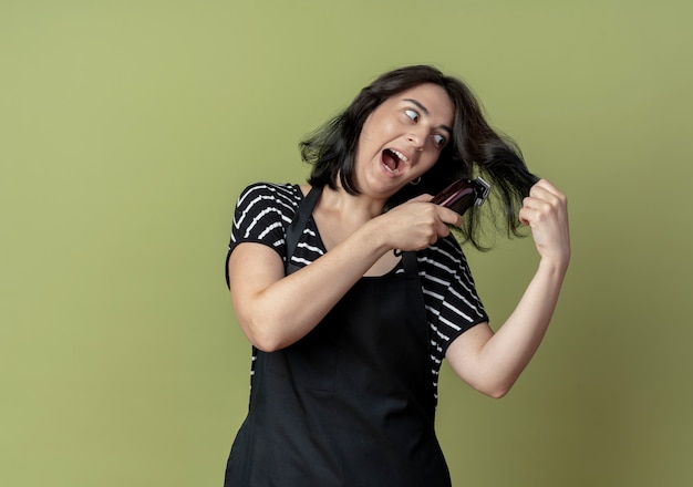 Foto gratuita peluquería mujer hermosa joven en delantal sosteniendo la máquina de corte de pelo que va a cortar su pelo muy emocional de pie sobre la pared