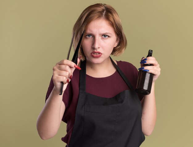 Peluquería de mujer hermosa joven en delantal sosteniendo pinza para el cabello botella de spray y peine mirando al frente con cara seria de pie sobre la pared verde