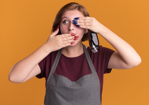 Peluquería de mujer hermosa joven en delantal sosteniendo recortadora cubriendo un ojo y boca con las manos de pie sobre la pared naranja