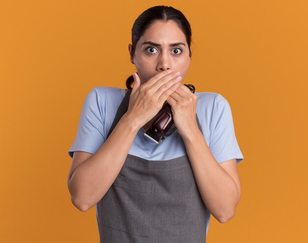 Peluquería de mujer hermosa joven en delantal sosteniendo recortadora mirando al frente siendo sorprendido cubriendo la boca con las manos sobre la pared naranja