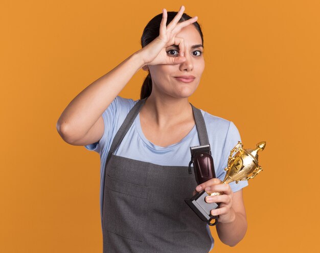 Peluquería de mujer hermosa joven en delantal sosteniendo recortadora y trofeo de oro mirando al frente sonriendo mostrando signo ok de pie sobre la pared naranja