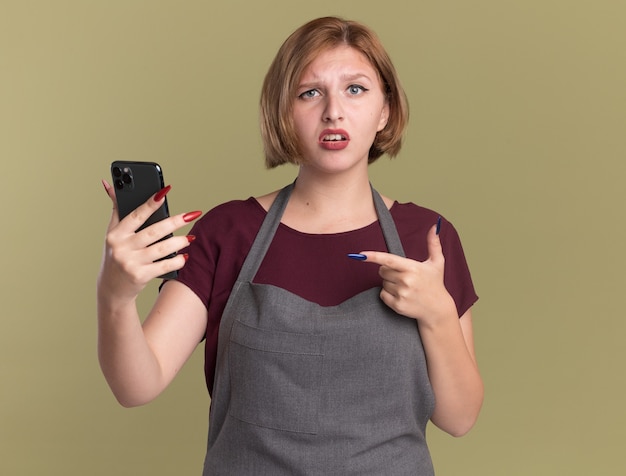 Peluquería de mujer hermosa joven en delantal sosteniendo smartphone apuntando con el dedo índice a confundirse de pie sobre la pared verde
