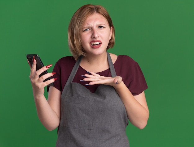 Peluquería de mujer hermosa joven en delantal sosteniendo smartphone mirando al frente confundido y disgustado de pie sobre la pared verde