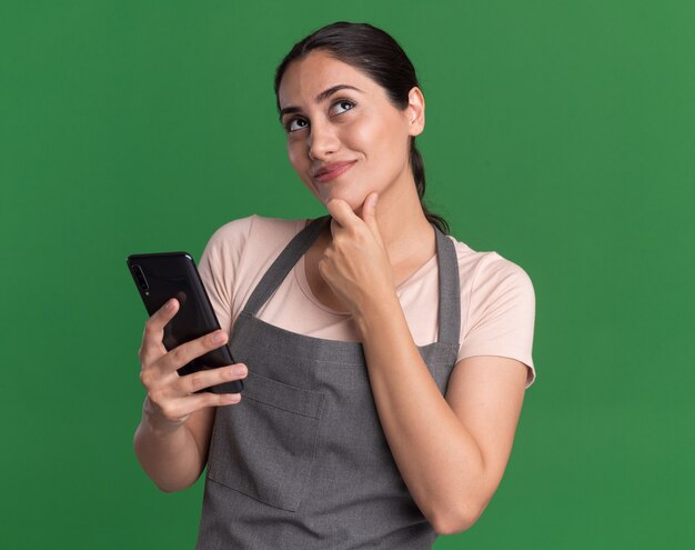 Peluquería de mujer hermosa joven en delantal sosteniendo smartphone mirando hacia arriba con expresión pensativa pensando y sonriendo de pie sobre la pared verde