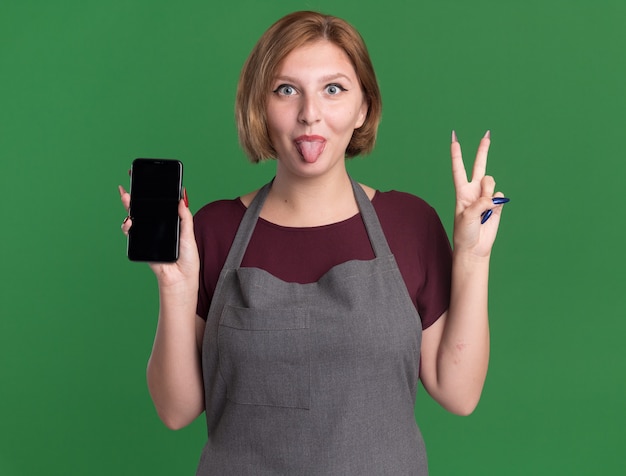 Peluquería de mujer hermosa joven en delantal sosteniendo smartphone mostrando v-sign sacando la lengua feliz y positivo de pie sobre la pared verde