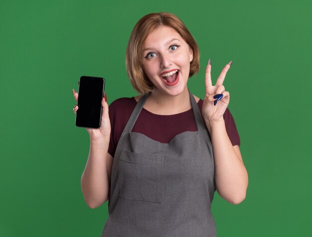 Peluquería de mujer hermosa joven en delantal sosteniendo smartphone mostrando v-sign sonriendo alegremente de pie sobre la pared verde