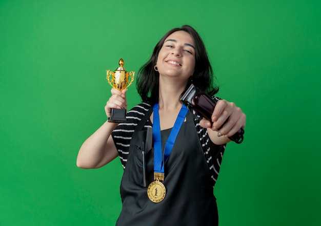 Foto gratuita peluquería mujer hermosa joven en delantal con trofeo y recortador de pie feliz y emocionado sobre la pared verde