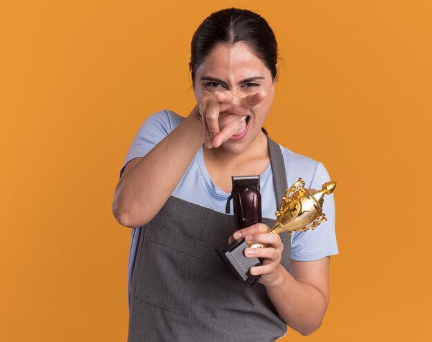 Peluquería de mujer hermosa joven divertida y alegre en delantal sosteniendo recortadora y trofeo de oro apuntando con los dedos en la parte delantera de pie sobre pared naranja