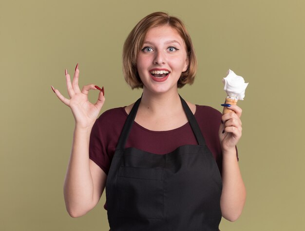 Peluquería de mujer hermosa joven feliz en delantal sosteniendo brocha de afeitar con espuma de afeitar mirando al frente sonriendo mostrando signo ok de pie sobre la pared verde