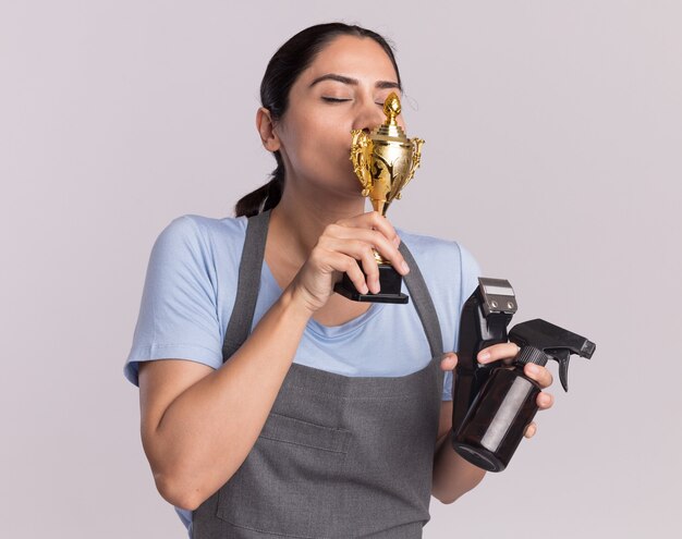 Peluquería de mujer hermosa joven feliz en delantal sosteniendo la máquina recortadora de trofeos de oro con botella de spray besando su trofeo de pie sobre la pared blanca