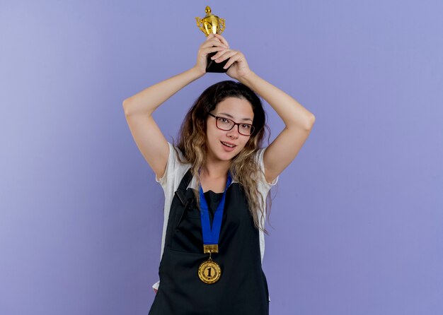 Peluquería profesional joven mujer en delantal con medalla de oro alrededor del cuello sosteniendo el trofeo sobre la cabeza feliz y positiva sonriendo de pie sobre la pared azul