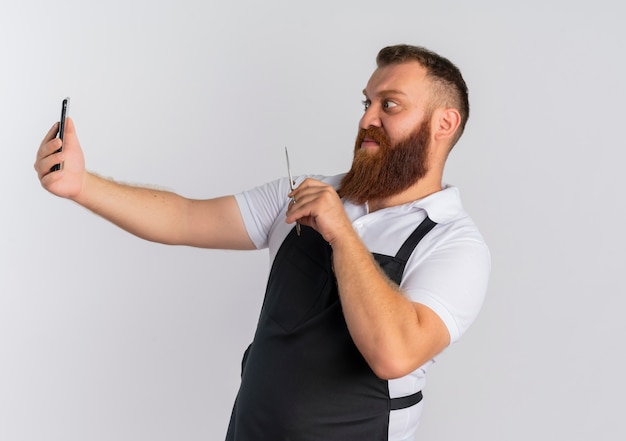 Peluquero barbero profesional en delantal con smartphone y tijeras, mirando confundido y molesto parado sobre la pared blanca