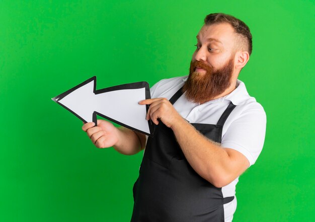 Peluquero barbudo profesional en delantal con gran cartel de flecha apuntando hacia la izquierda de pie sobre la pared verde