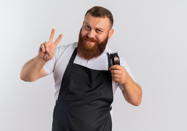 Foto gratuita peluquero barbudo profesional en delantal sosteniendo la máquina cortadora de cabello mostrando el signo de la victoria o el número dos sonriendo de pie sobre la pared blanca