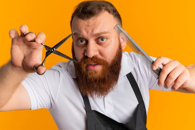 Peluquero barbudo profesional en delantal con tijeras y cepillo para el cabello que va a cortar de pie sobre la pared naranja