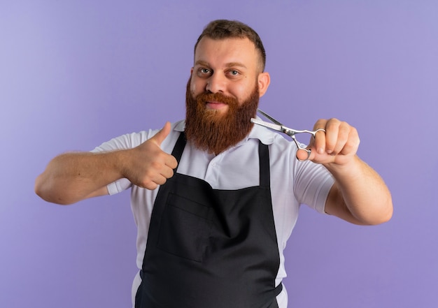 Peluquero barbudo profesional hombre en delantal sosteniendo tijeras sonriendo alegremente mostrando los pulgares para arriba de pie sobre la pared púrpura