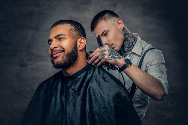 Un peluquero caucásico tatuado cortando la barba a un hombre negro con estilo.